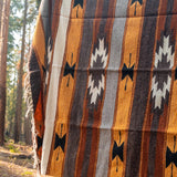A woven southwestern-style blanket with vertical stripes and geometric motifs in mustard yellow, brown, gray, black, and off-white, draped outdoors in a forest setting.