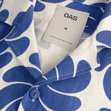 "close up on collar and necl tag blue cartoonish flower or palm leaves on a cream button up linen shirt"