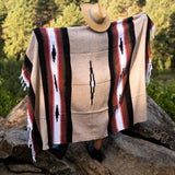 A woven poncho blanket with Southwestern geometric patterns in beige, black, red, and white, draped over a person sitting on rocks outdoors with a tan hat resting on their head.