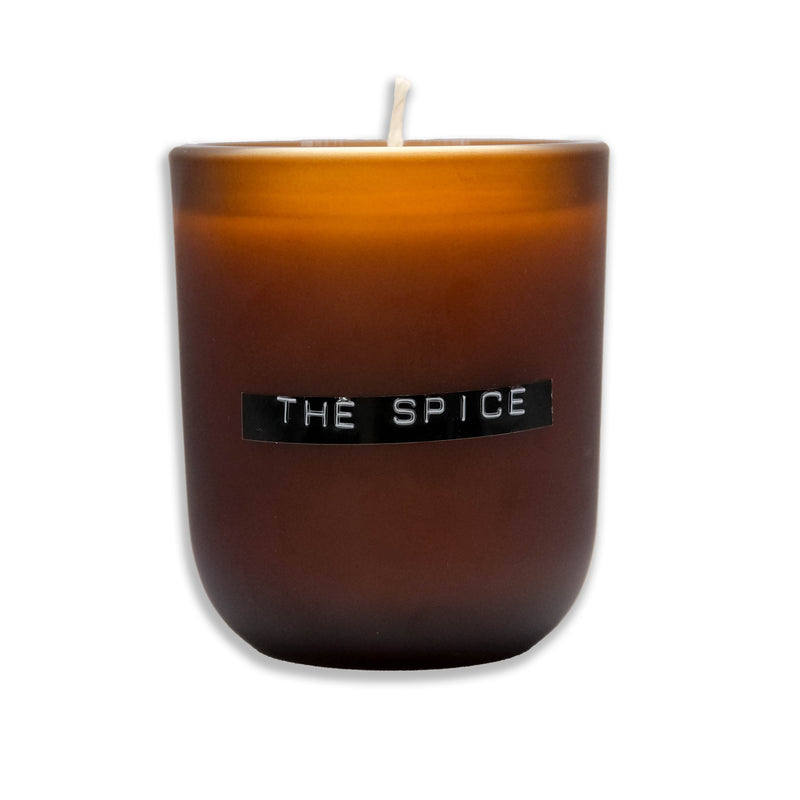 A brown soy wax candle with a label that reads 'THE SPICE' on the front, with a visible wick on top.