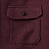 Close up of chest pocket on a dark burgundy shirt with a flap that is secured by a black buttons. There is a subtle cross-hatching texture on the fabric of the shirt. 