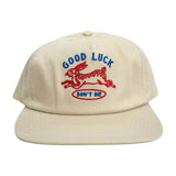 Ecru colored baseball hat facing forward with blue and red embroidery of a leaping rabbit with text [Good Luck Don't Die]