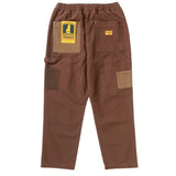 Back of a pair of straight leg canvas pants in shades of color blocked brown. There is a hammer loop on the side of the wearer's left pant leg. There are two back pockets. The left pocket has a yellow and black cardboard tag with a cartoon waiter and the text [Service Works] on it and the right has a small orange fabric patch sewn on it with the Service Works logo. 